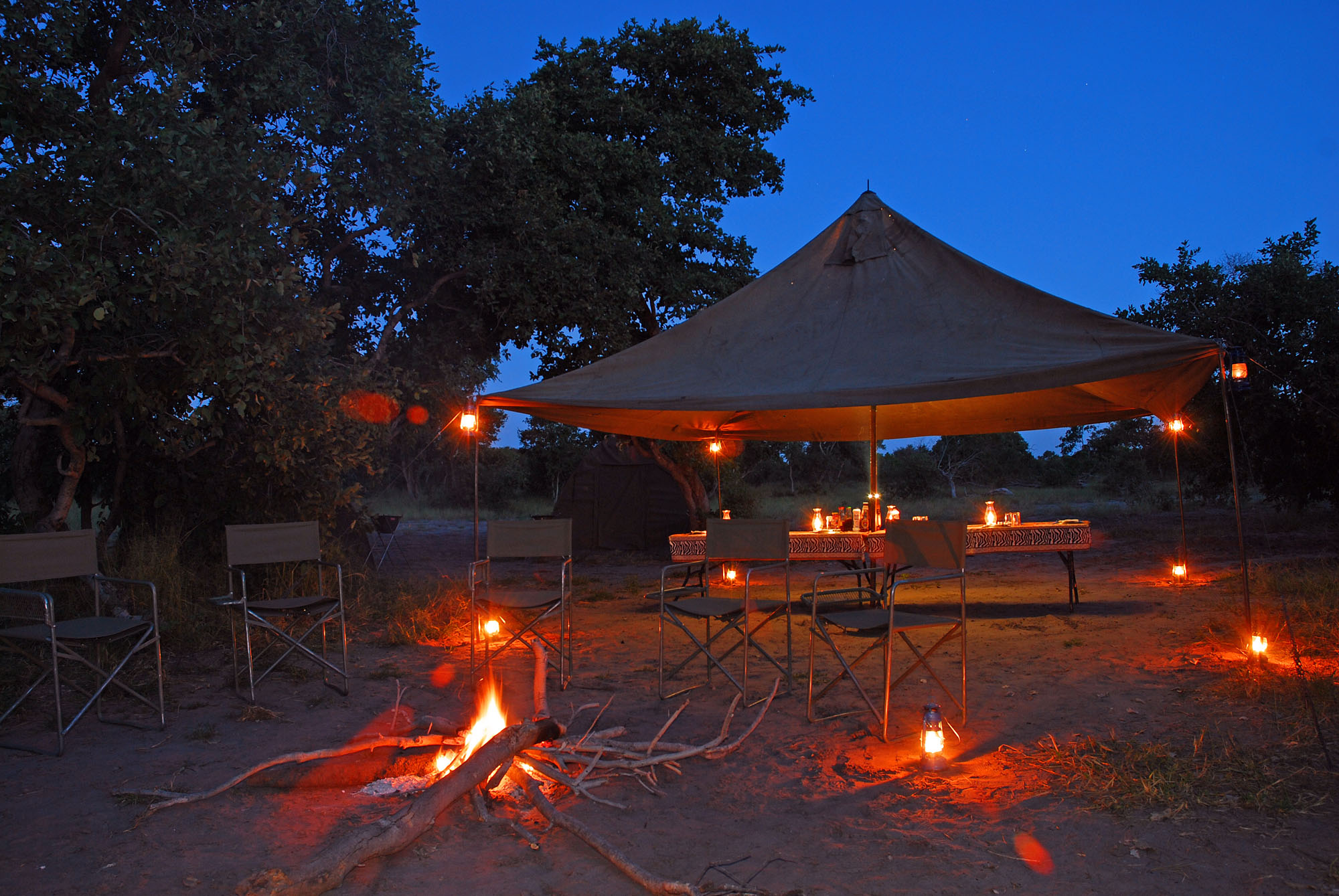 Gemütliches Safari-Camp in Botswana bei Einbruch der Dunkelheit mit Lagerfeuer, Laternen und überdachtem Essbereich unter freiem Himmel. Authentisches Wildnis-Camp inmitten der afrikanischen Natur während einer Safari-Rundreise durch Botswana.
