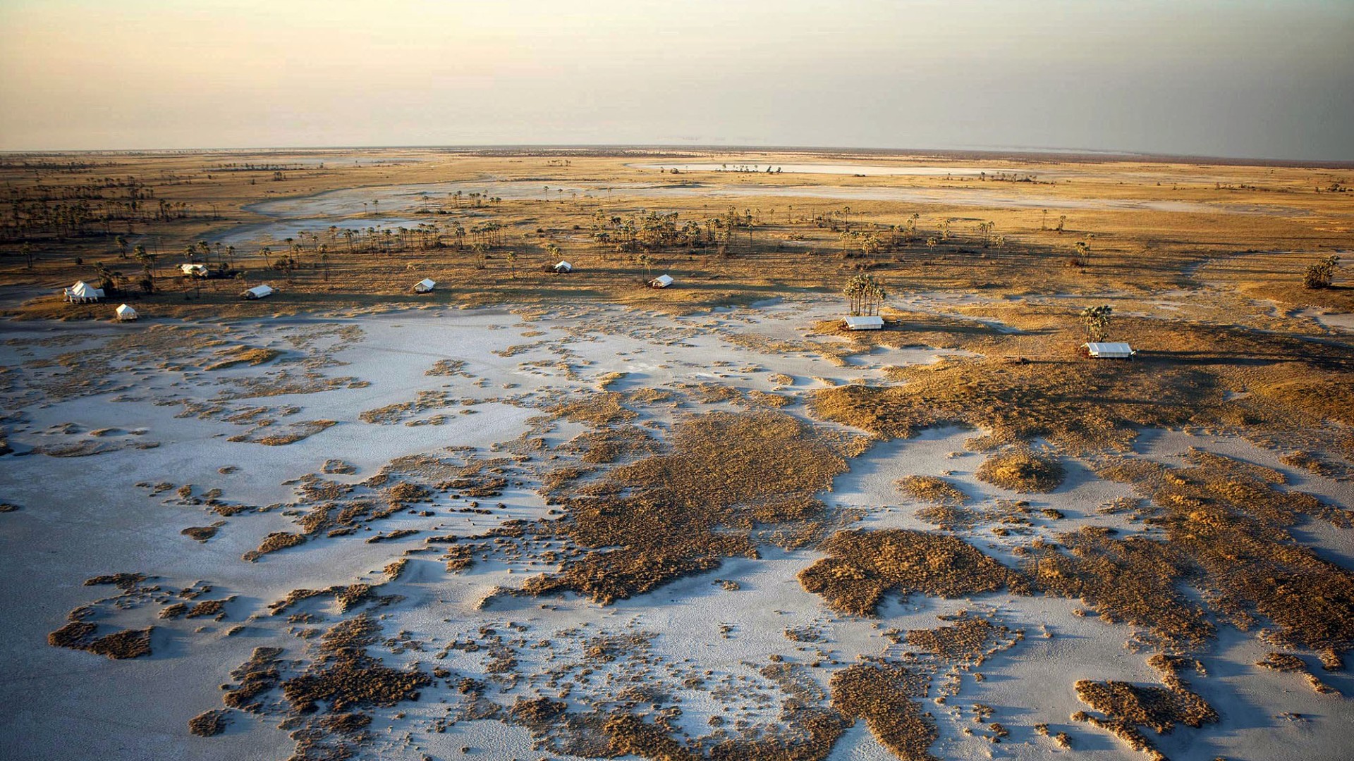 Luftaufnahme des Okavango-Deltas in Botswana mit weitläufigen Wasserflächen, Grasinseln und abgelegenen Safari-Camps inmitten einer einzigartigen Wildnislandschaft.