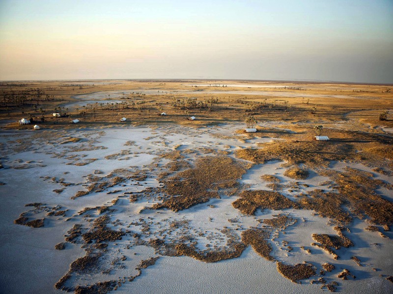 Luftaufnahme des Okavango-Deltas in Botswana mit weitläufigen Wasserflächen, Grasinseln und abgelegenen Safari-Camps inmitten einer einzigartigen Wildnislandschaft.