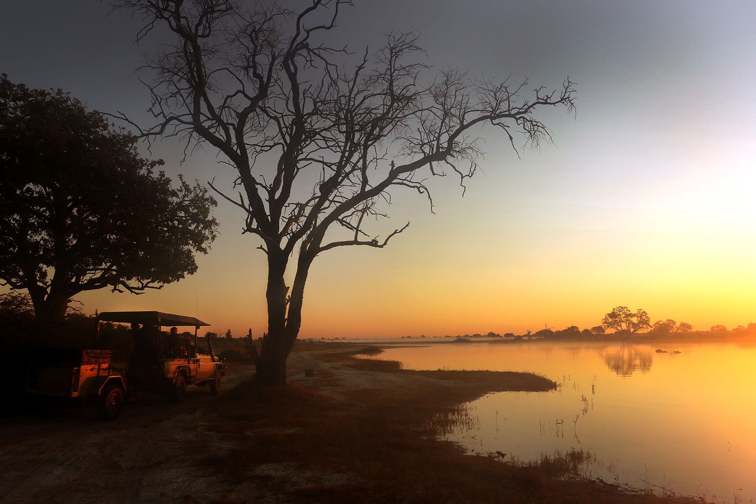 Safari-Fahrzeug am Ufer eines Wasserarms im Okavango-Delta bei Sonnenaufgang, Botswana.