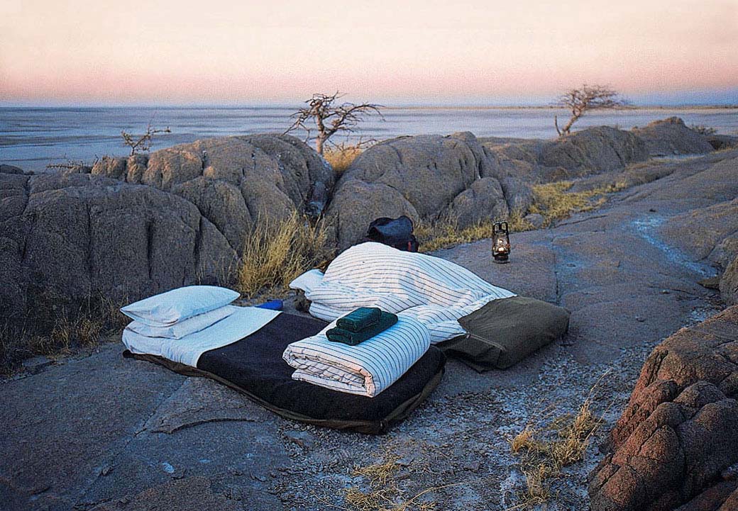 Bett unter freiem Himmel auf den Granitfelsen von Kubu Island in Botswana mit Blick über die Weite der Makgadikgadi-Salzpfannen bei Abenddämmerung.