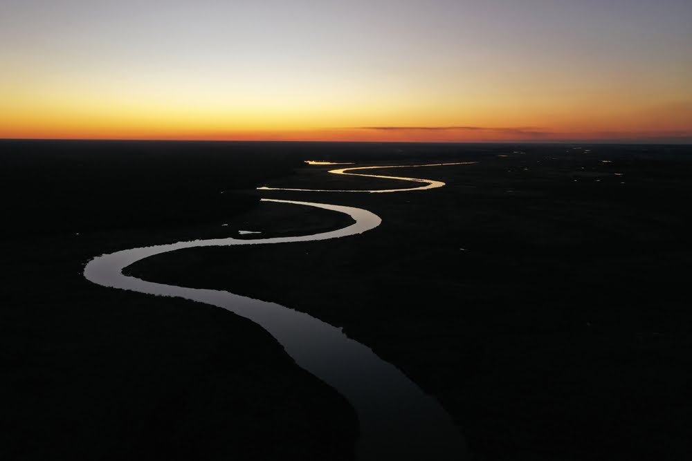 Luftaufnahme eines gewundenen Flusslaufs im Abendlicht der botswanischen Wildnis, der sich durch die weite Landschaft des Okavango-Systems schlängelt.