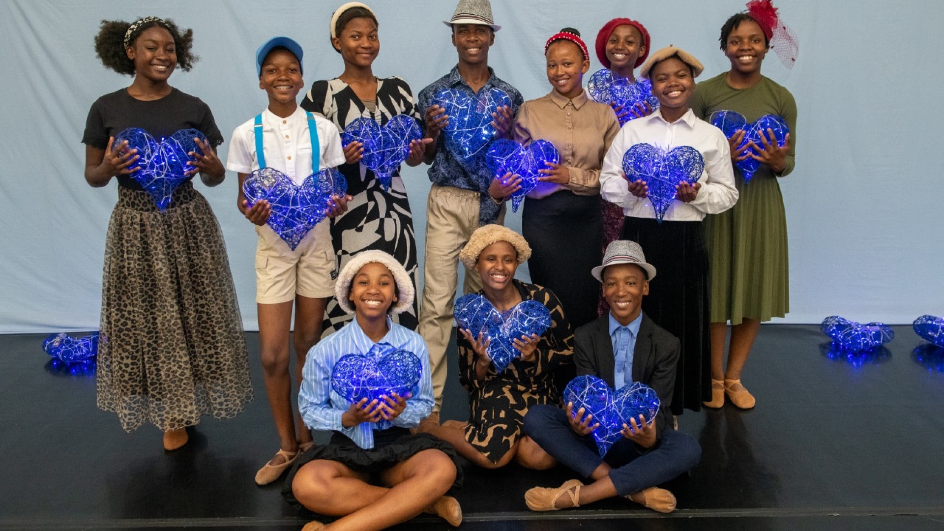 Eine Gruppe junger Menschen präsentiert im Rahmen eines afrikanischen Community-Projekts kunstvoll gestaltete, leuchtend blaue Herzskulpturen. Die Jugendlichen stehen und sitzen gemeinsam auf einer Bühne und symbolisieren Zusammenhalt, Kreativität, Bildun