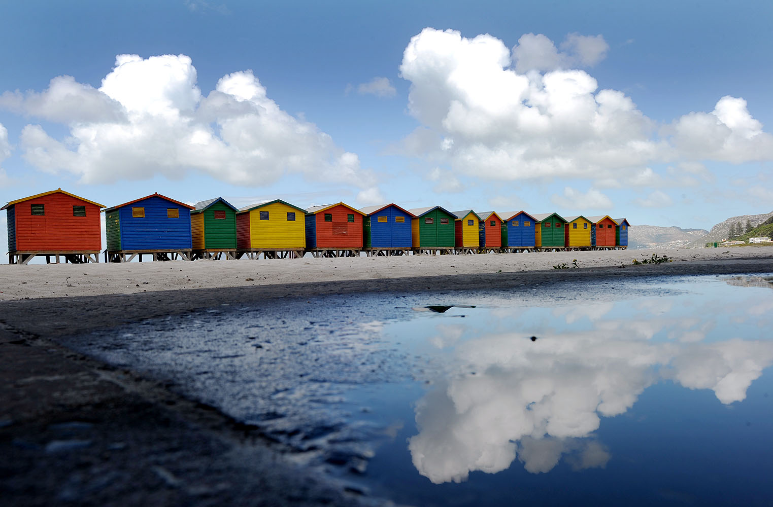 Farbenfrohe Strandhütten am Muizenberg Beach bei Kapstadt spiegeln sich nach einem Regenschauer in einer Wasserpfütze.