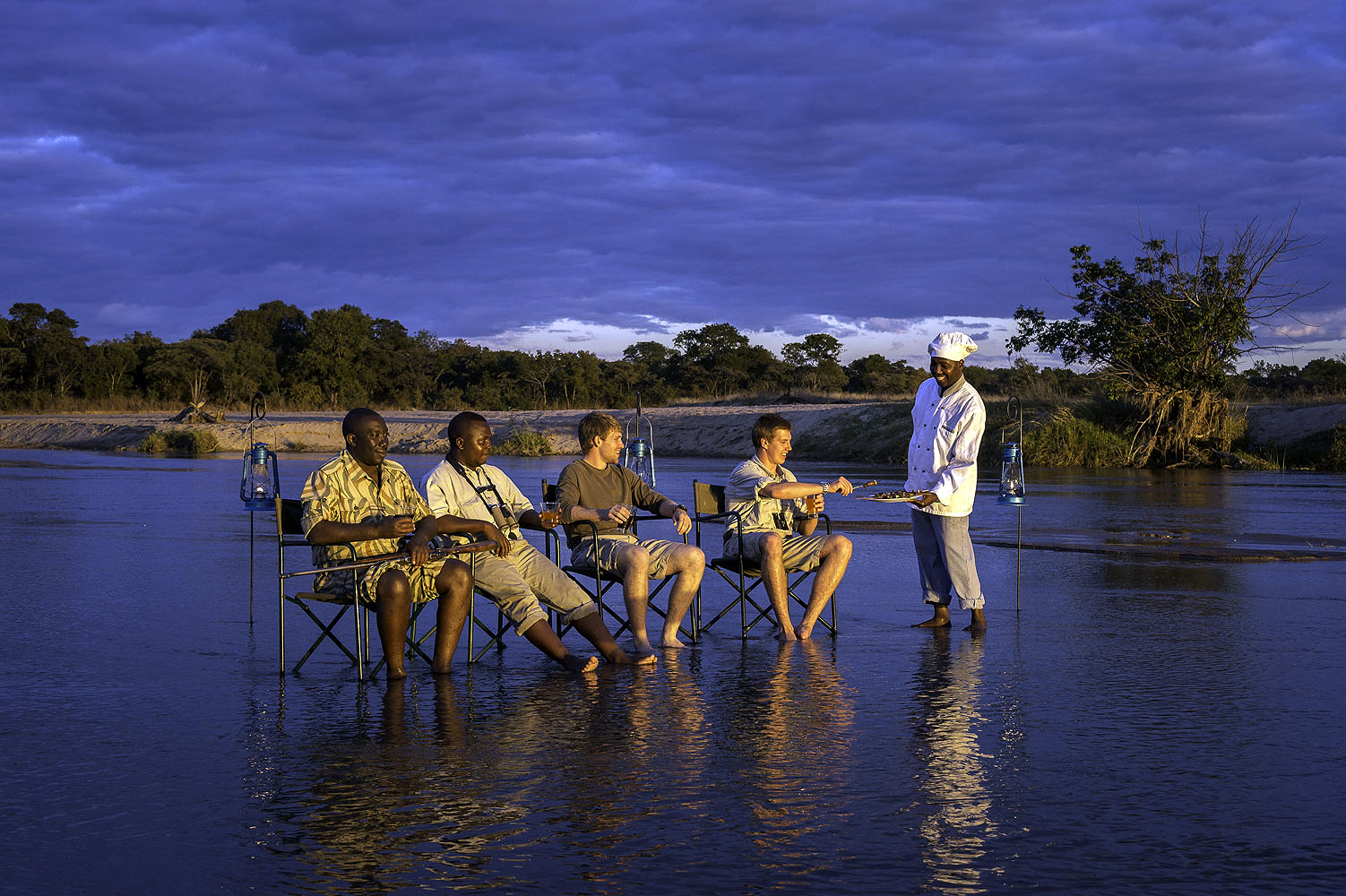Safari-Gäste genießen ein exklusives Abendessen mitten im flachen Fluss, serviert von einem Koch bei Sonnenuntergang in Afrika.